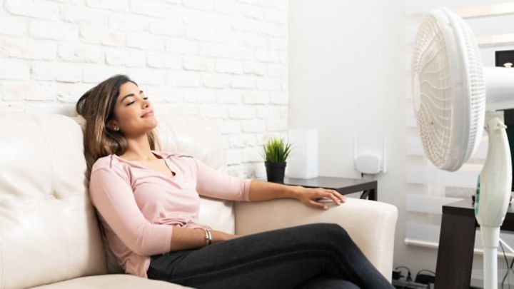 © stock.adobe.com/AntonioDiaz Eine Frau liegt entspannt auf dem Sofa vor einem Ventilator.