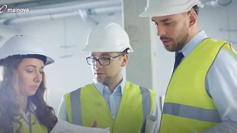 Drei Personen mit Schutzhelmen und Warnwesten besprechen Baupläne auf einer Baustelle.