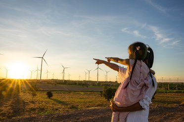 © Quelle: https://www.istockphoto.com/nensuria Mutter mit Tochter auf dem Arm schauen zu einem Windpark bei Sonnenaufgang