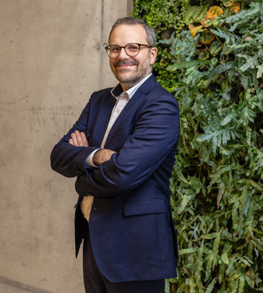 © Mainova Ein lächelnder Moritz Möller mit Brille, gekreuzten Armen und dunklem Anzug steht vor einer Wand mit vertikaler Begrünung.