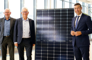 © Mainova Herr Giehl mit zwei Herren steht lächelnd neben einem großen Solarmodul in einem hellen Büro.