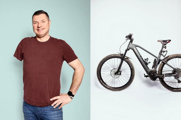 © Ramon Haindl Mann in braunem Shirt lächelt, Hand auf Hüfte; daneben E-Bike vor weißem Hintergrund.