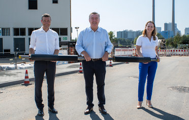 Zwei Männer und eine Frau stehen vor einem Kraftwerk mit einem Fernwärmerohr in den Händen