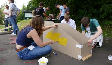 © Mainova AG Junge Menschen sitzen auf der Erde und bemalen eine Seifenkiste aus Karton