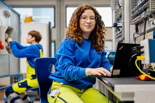 © Stilmanöver/RamonHaindl Junge Frau in Arbeitskleidung sitzt am Laptop in einer Werkstatt, im Hintergrund arbeitet ein Kollege an einer Anlage.