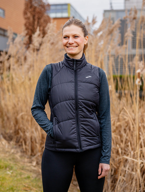 © Stilmanöver/RamonHaindl Frau in Sportkleidung steht lächelnd vor hohem Schilfgras, moderne Gebäude im Hintergrund.
