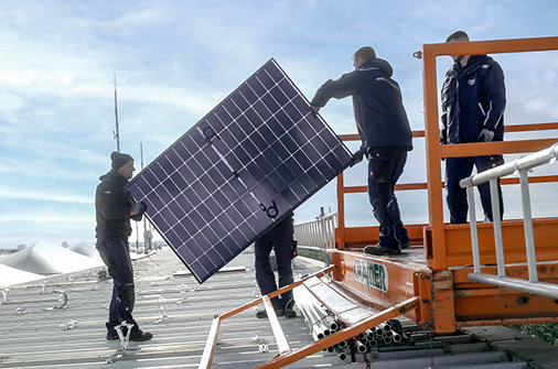 Vier Personen installieren ein Solarpanel auf einem Dach.