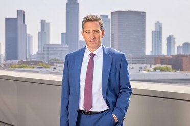© mainova/JoachimStorch Ein Mann im blauen Anzug mit roter Krawatte steht auf einer Terrasse vor der Skyline von Frankfurt am Main.