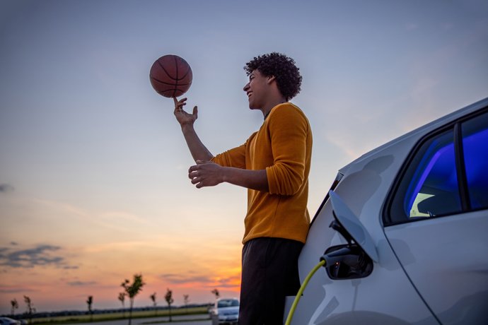 Mann dreht Basketball auf Fingerspitze und steht vor einem Sonnenuntergang angelehnt am ladenden Auto