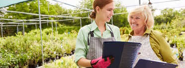Zwei Personen in einer Gärtnerei betrachten Ordner, umgeben von grünen Pflanzen in Töpfen.