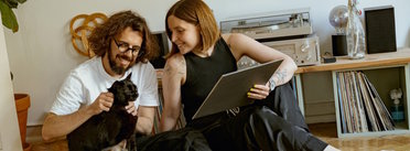 Zwei Personen sitzen auf dem Boden, eine hält eine Schallplatte, die andere streichelt eine schwarze Katze.