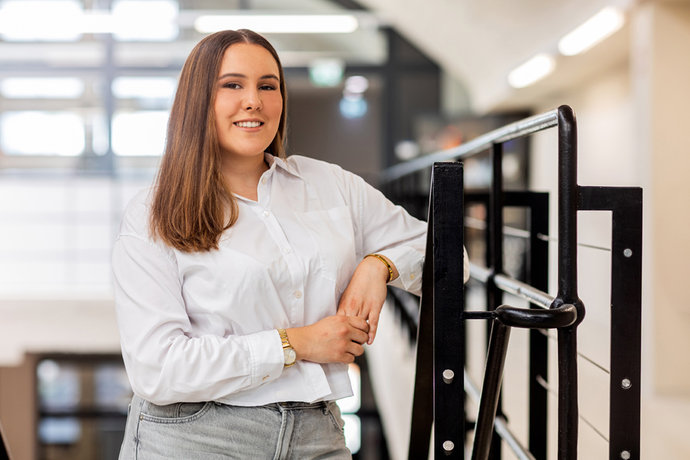 Susanna Scholz mit weißer Bluse und langer Hose steht lächelnd an einem Geländer im Innenraum.