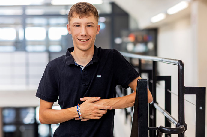 Lukas Engel im schwarzen Polohemd lehnt lächelnd an einem Geländer in einem modernen Gebäude.