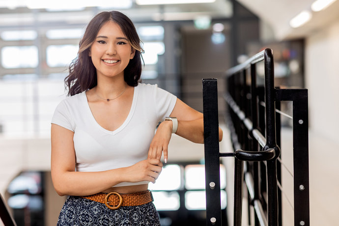 Eliza Maksatbekova mit weißem Shirt und gemustertem Rock lehnt lächelnd an einem schwarzen Geländer.