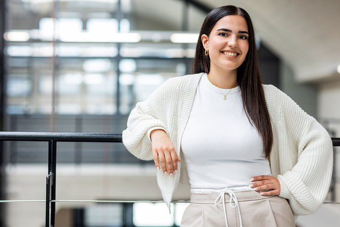 Junge Frau lehnt lächelnd an Geländer in modernem Gebäude, trägt helle Kleidung und dezenten Schmuck.