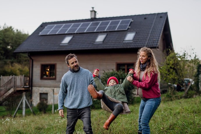 Personen spielen mit einem Kind vor einem Wohnhaus mit Solaranlage auf dem Dach in ländlicher Umgebung.