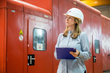 Eine Person mit Helm und Klemmbrett steht vor einer roten Industrietür.