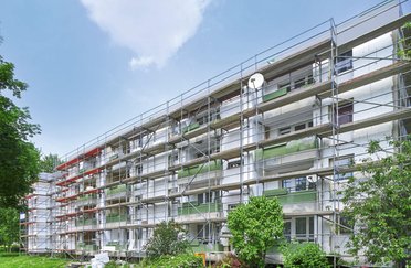 Ein mehrstöckiges Gebäude mit Gerüsten an der Fassade, umgeben von Bäumen und Grünflächen.