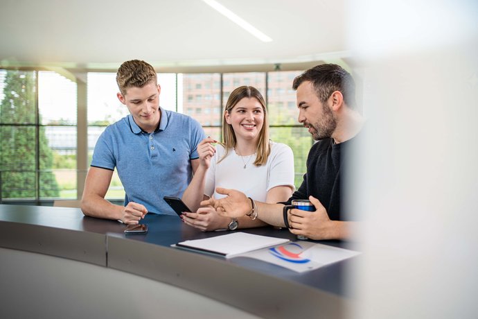Duale Studierende stehen am Tisch und schauen sich den Display eines Smartphone an