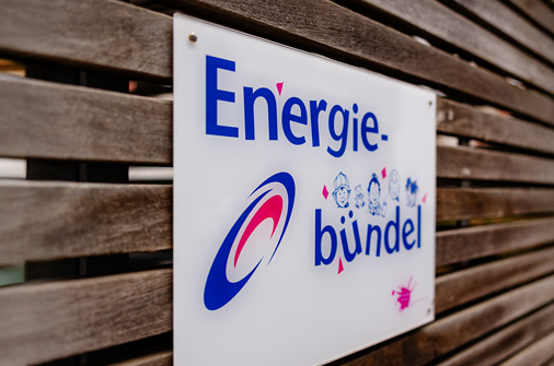 Schild mit der Aufschrift Energiebündel samt Logo und Kinderzeichnungen, montiert an einer Holzlattenwand.