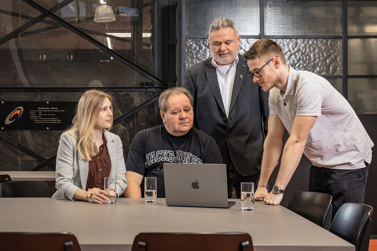 © Stilmanöver/RamonHaindl Vier Personen schauen gemeinsam auf einen Laptop, sie sitzen und stehen um einen Tisch mit Wassergläsern.