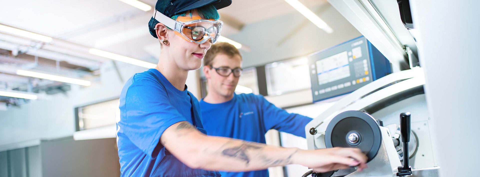 © Mainova/AndreFormer Junge Frau steht an Drehmaschine und der Ausbilder schaut der Arbeit zu.