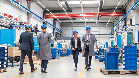 © iStock.com/vm Geschäftsmänner und Geschäftsfrauen laufen durch eine hell beleuchtete Produktionshalle