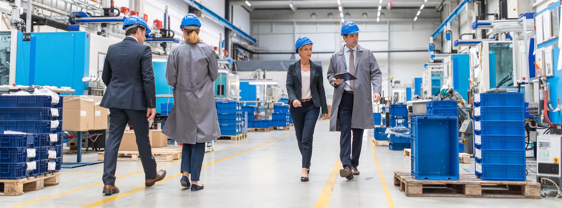 © iStock.com/vm Geschäftsmänner und Geschäftsfrauen laufen durch eine hell beleuchtete Produktionshalle