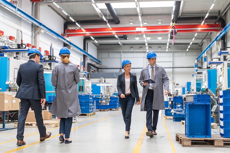 © iStock.com/vm Geschäftsmänner und Geschäftsfrauen laufen durch eine hell beleuchtete Produktionshalle