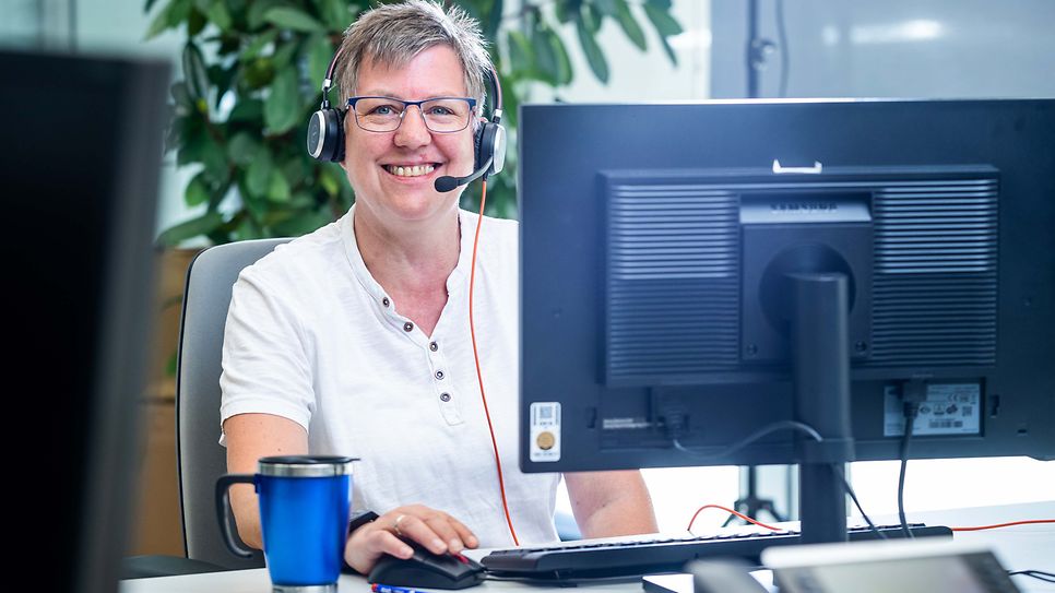 © Mainova/AndreFormer Bettina Rakowksi sitzt mit einen Headset am Computer und blickt über den Schreibtisch in die Kamera
