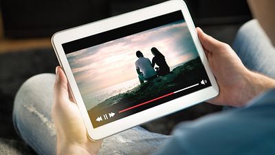 Tablet in den Händen einer Person, zeigt ein Video, in dem ein Paar am Meer sitzt