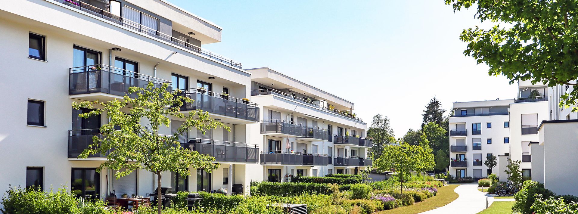 Moderne Wohnanlage mit mehreren weißen Gebäuden, Balkonen und gepflegten Grünflächen bei sonnigem Wetter.