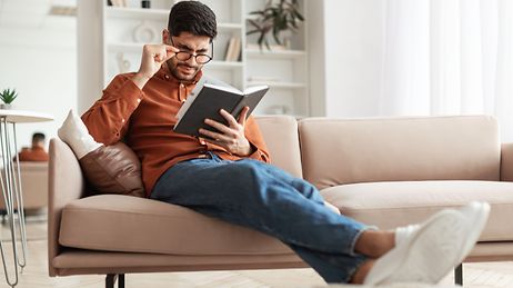 © Quelle: https://www.istockphoto.com/Prostock-Studio Junger Mann sitzt auf dem Sofa und liest ein Buch.