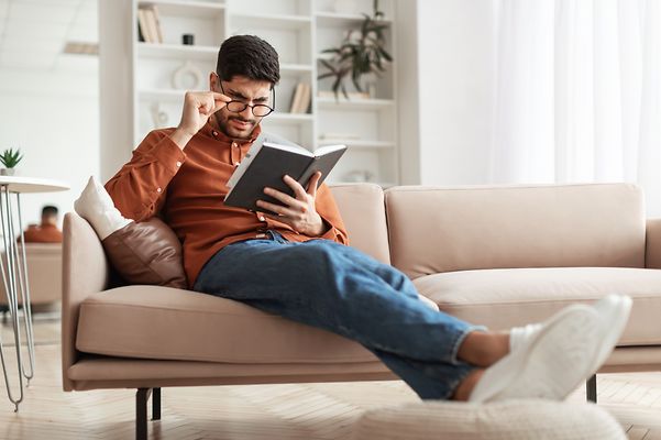 Junger Mann sitzt auf dem Sofa und liest ein Buch.