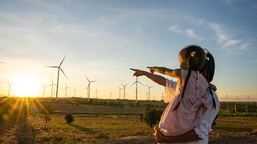 © Quelle: https://www.istockphoto.com/nensuria Mutter mit Tochter auf dem Arm schauen zu einem Windpark bei Sonnenaufgang