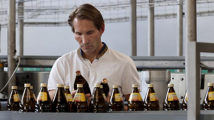 Ein Mann in einem weißen Hemd steht hinter einer Reihe von braunen Flaschen auf einem Förderband in einer Fabrik.