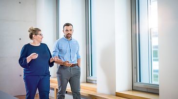 © Mainova/AndreFormer Frau und Mann laufen vor Fensterwand und sprechen miteinander