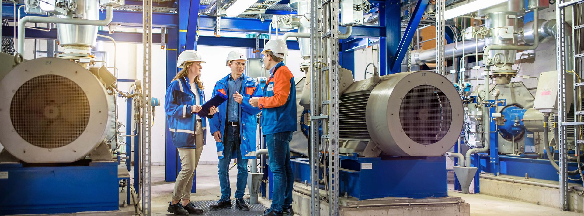 © Mainova/AndreFormer Drei Trainees mit Helm besprechen sich in einer Maschinenhalle