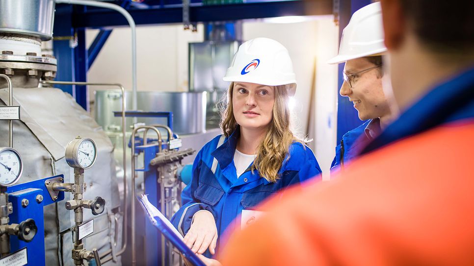 © Mainova/AndreFormer Junge Ingenieurin mit Helm hört ihren Kollegen vor einer Maschine zu