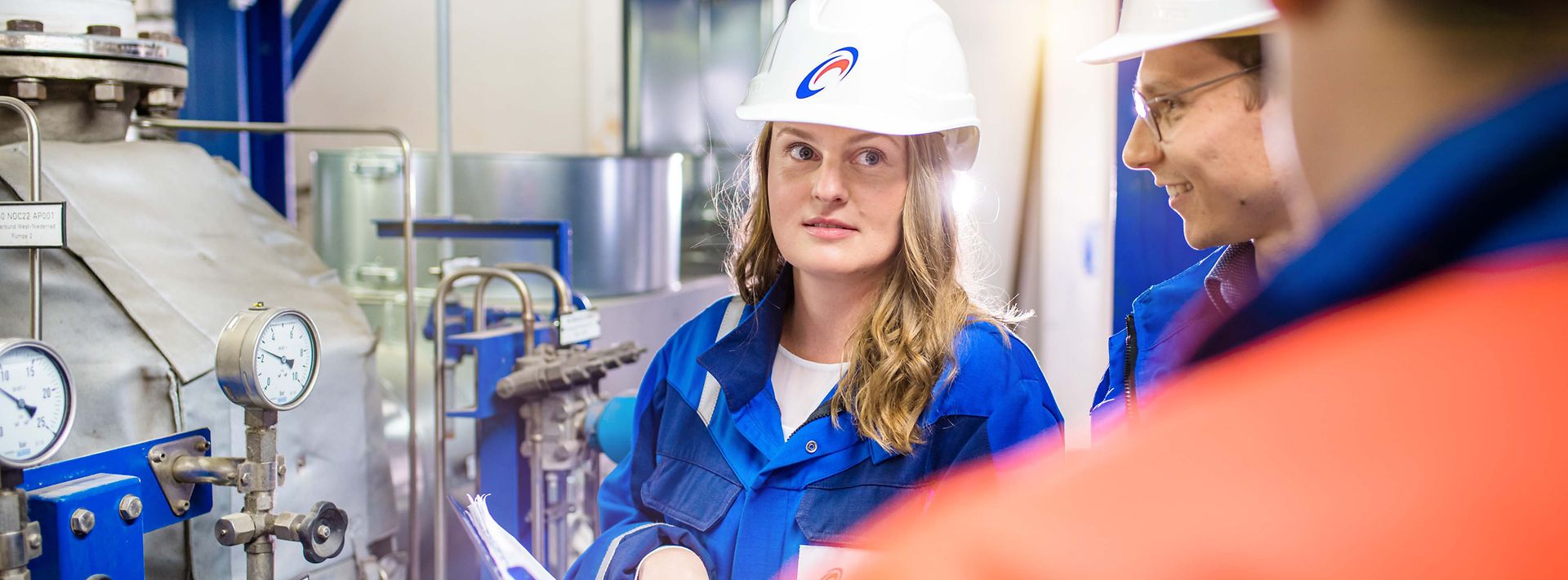 Junge Ingenieurin mit Helm hört ihren Kollegen vor einer Maschine zu