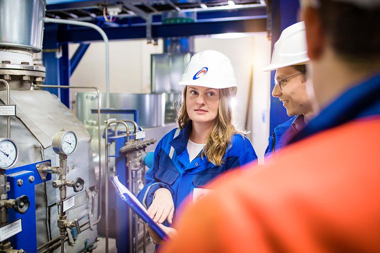© Mainova/AndreFormer Junge Ingenieurin mit Helm hört ihren Kollegen vor einer Maschine zu