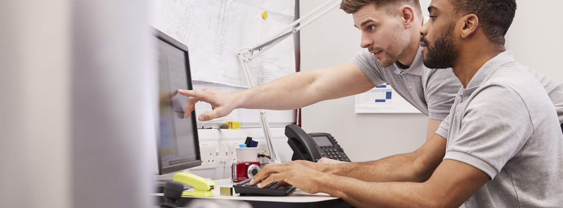 Ein Mann zeigt einem am Computer sitzenden Arbeitskollegen etwas auf dem Computer-Bildschirm.