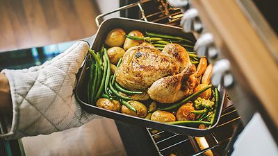 Eine Hand mit Ofenhandschuh holt eine Bratform mit Gänsebraten, Kartoffeln und Bohnen aus dem Ofen
