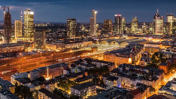 Beleuchtetes Frankfurt in der Nacht: Skyline im Hintergrund, Bahnhof und Zuggleise in der Mitte und Wohnquartier vorne.