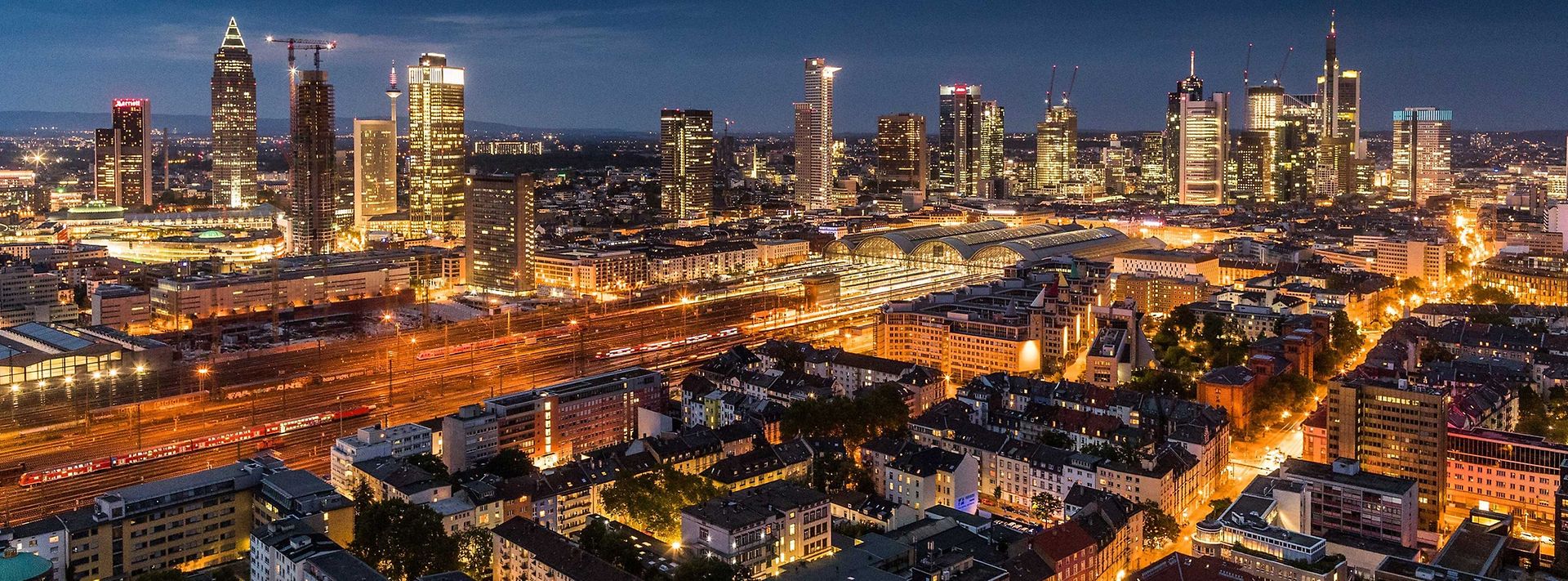 Beleuchtetes Frankfurt in der Nacht: Skyline im Hintergrund, Bahnhof und Zuggleise in der Mitte und Wohnquartier vorne.