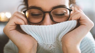 Frau mit Brille zieht weißen Rollkragen über Mund und Nase