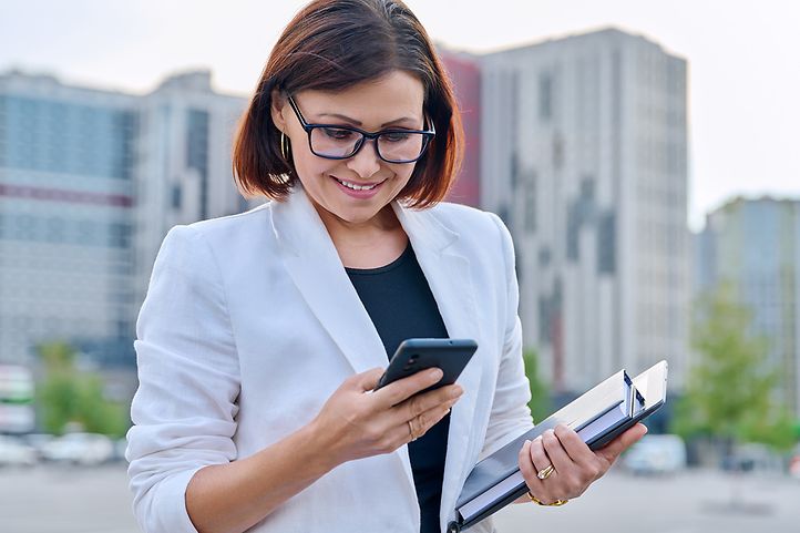 Eine Frau in einem weißen Blazer hält ein Smartphone und einen Ordner, während sie draußen steht.