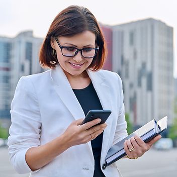 Eine Frau in einem weißen Blazer hält ein Smartphone und einen Ordner, während sie draußen steht.