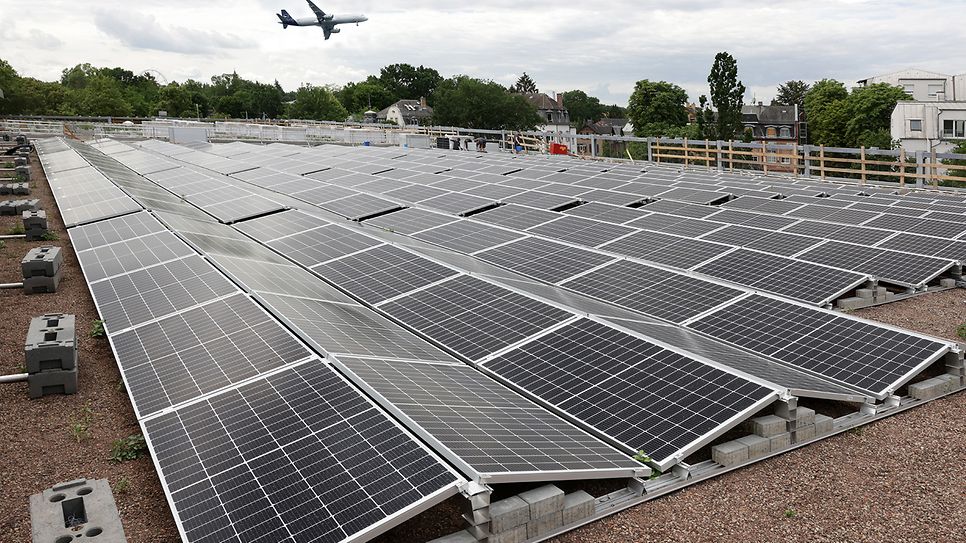 © Mainova Dach mit vielen Solarpaneelen, im Hintergrund fliegt ein Flugzeug über grüne Landschaft.
