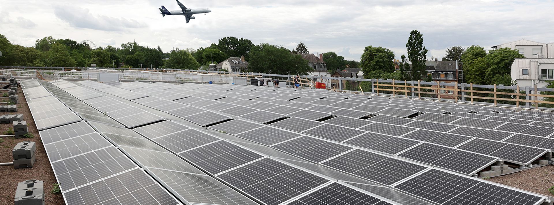 © Mainova Dach mit vielen Solarpaneelen, im Hintergrund fliegt ein Flugzeug über grüne Landschaft.